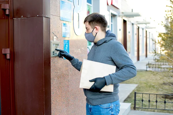Courier delivers the parcel to the door, rings the intercom. Men in latex gloves and medical mask contactless delivers a package with products, goods to the client during the quarantine period.