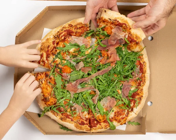 Eating traditional italian pizza with tomatoes, prosciutto, arugula and mozzarella cheese top view. A child and a man eating pizza from delivery carton box