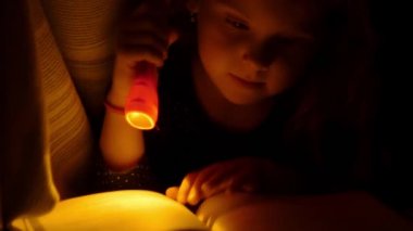Kız yatak örtüsü altında bir el feneri ile kitap okuyorum. Yatakta okuma çocuk. Yakın çekim