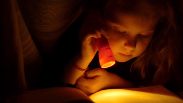 Kız el feneri ile yorganın altında okuma. Yatakta çocuk okuma. Küçük kız yatakta kitap okuyarak kapatın — Stok video
