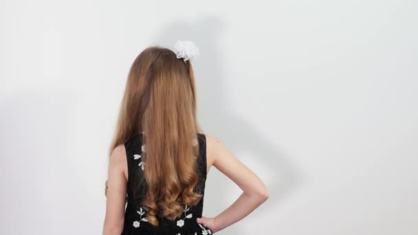 Beautiful girl with long hair turning around and winks. Charming little girl stands in front of white background, turns around and winks. — Stock Video