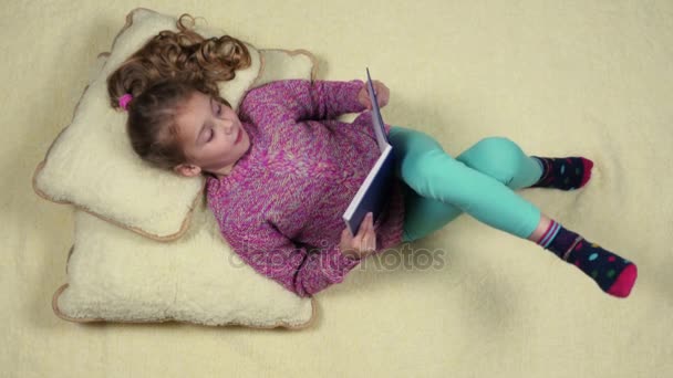 Vicces kis lány feküdt a kanapén, egy könyvet olvas. Szemközti nézet — Stock videók