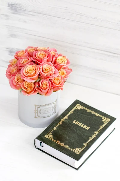Pink roses in a round box and the Bible on white wooden background.