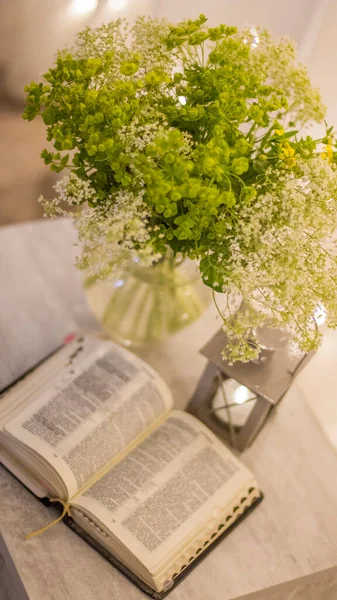 Composition: an open Bible, a bouquet of wildflowers in a vase and a decorative flashlight with a candle on the nightstand against the background of burning lights.