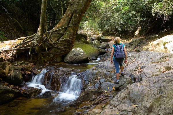 Uma menina com uma mochila fica perto de uma pequena cachoeira pitoresca — Fotografia de Stock