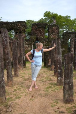 A cute, natural blonde with no shoes stands next to the ancient 