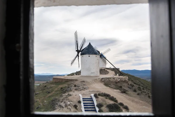 White old windmill on the hill near Consuegra (Castilla La Mancha, Spain), a symbol of region and journeys of Don Quixote (Alonso Quijano) and a town on cloudy day, a view through the window. — Stock Photo, Image