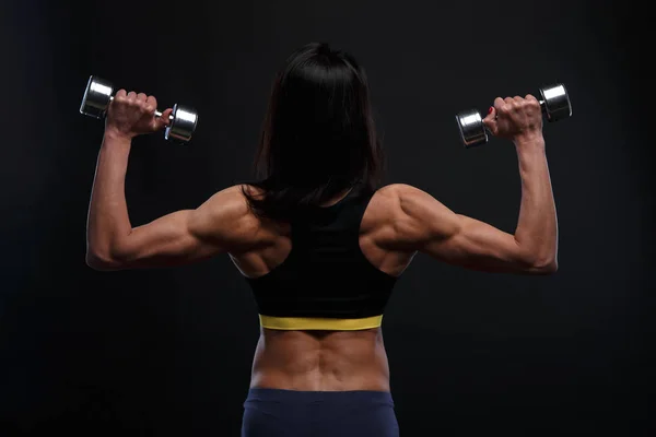 Achterzijde mooi atletische meisje werken met gewichten op een donkere achtergrond — Stockfoto