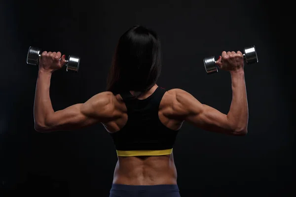 Achterzijde mooi atletische meisje werken met gewichten op een donkere achtergrond — Stockfoto