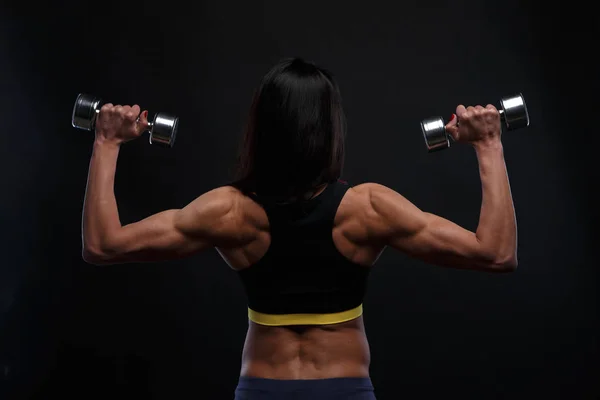 Espalda de hermosa chica atlética trabajando con pesas sobre un fondo oscuro —  Fotos de Stock