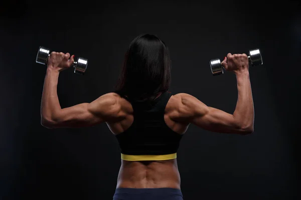 Achterzijde mooi atletische meisje werken met gewichten op een donkere achtergrond. — Stockfoto