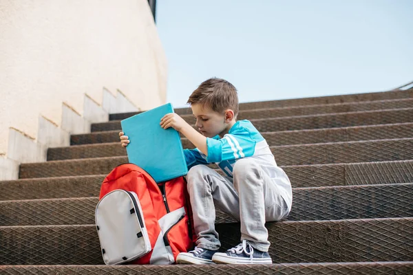 Jeune écolier assis devant une école et sortant un livre — Photo