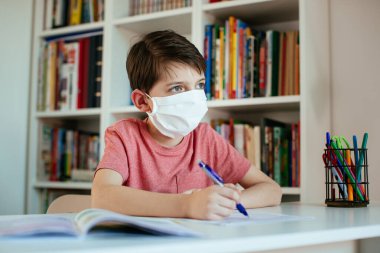 Koronavirüs salgını sırasında yüz maskesi takan çocuk evde kendi kendine çalışıyor. Ameliyat maskesi takan genç bir öğrenci okul ödevlerini kendi başına yapıyor..