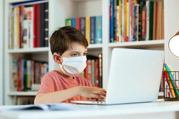 Yüz maskesi takan çocuk evde öğreniyor. Cerrahi maske takan genç öğrenci ödevini yapıyor..