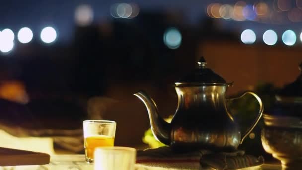 Maceta y vasos de metal tradicionales con té marroquí al vapor en la terraza de la azotea con vistas a la ciudad nocturna, medianoche, luz nocturna — Vídeo de stock