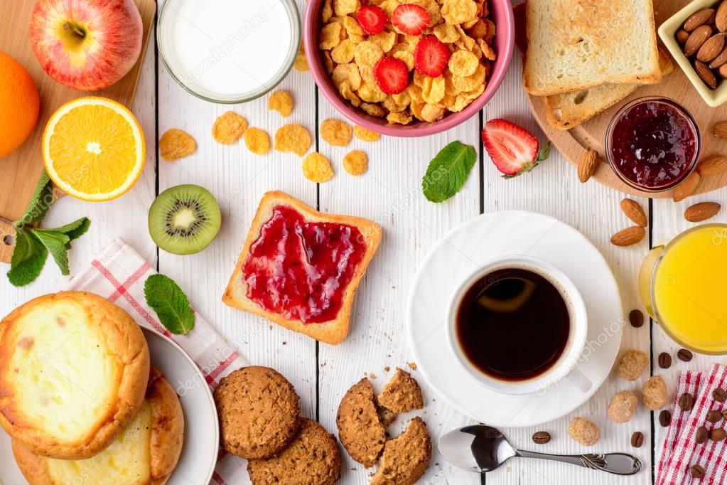 Breakfast table with healthy tasty ingredients. Coffee, toast, jam, cheesecakes, corn flakes, cookies, almonds, milk, orange juice and fruit on white wooden background.