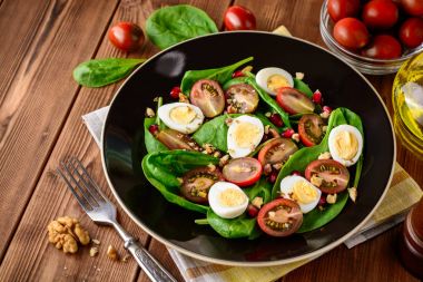 Ispanak, cherry domates, bıldırcın yumurtası, nar taneleri ve ceviz ahşap masa üzerinde siyah plaka ile taze sebze salatası.