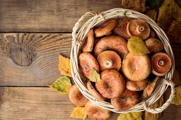 Ryzec pravý, běžně známý jako šafránem mléka čepici a borovice červená hub v proutěném koši na rustikální dřevěné pozadí s podzimní listí. — Stock fotografie