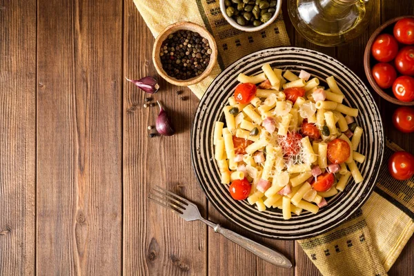 Makaron z boczkiem, czosnkiem, pomidorami, kaparami i parmezanem ser w płycie na ciemnym tle drewnianych. — Zdjęcie stockowe