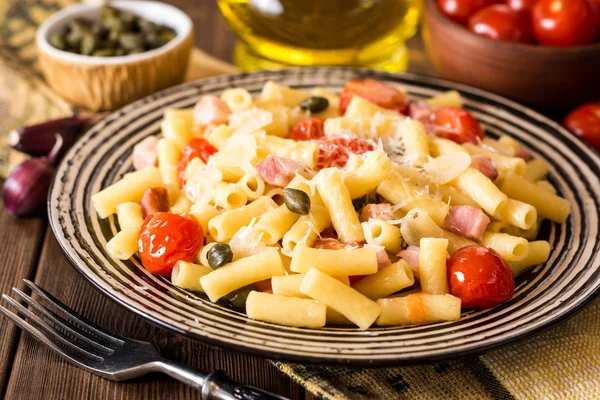 Pasta con tocino, ajo, tomates, alcaparras y queso parmesano en plato sobre fondo de madera oscura . — Foto de Stock