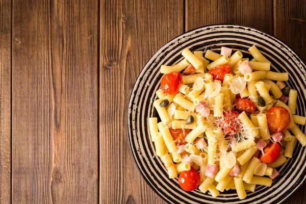 Pasta con tocino, ajo, tomates, alcaparras y queso parmesano en plato sobre fondo de madera oscura . — Foto de Stock