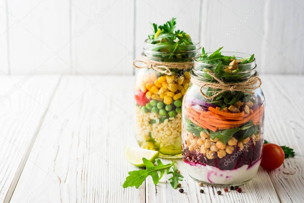Healthy homemade salads with chickpeas, bulgur and vegetables in mason jars on white wooden background.