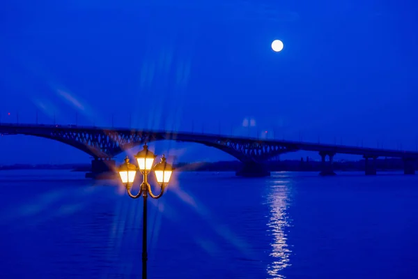 Lâmpada de rua luminosa em primeiro plano. No fundo a ponte sobre o rio. Passeio noturno ou noturno na cidade de Saratov, Rússia. O rio Volga. A lua cheia — Fotografia de Stock