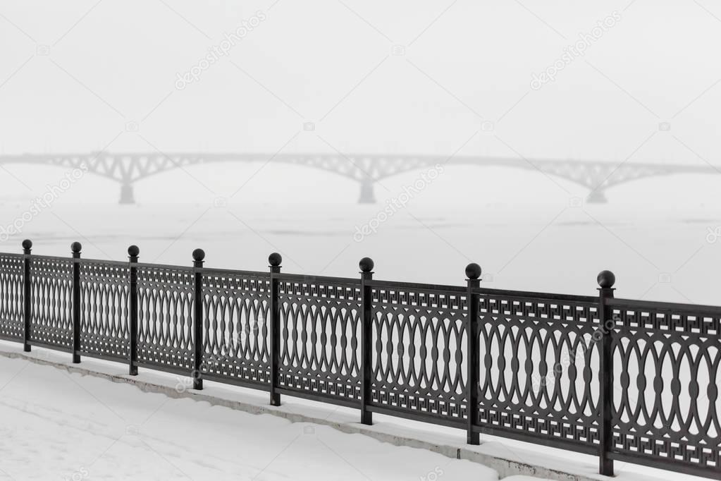 The metal fence on the waterfront in the city of Saratov, Russia. In the background is the road bridge across the Volga River. Winter cloudy day. Ice on the river.