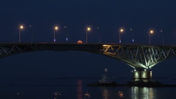 Повний місяць піднімається над мостом. Уповільнена. Автомобільного мосту між містами Саратов і Енгельс, Росія. Волги. Увечері вогні автомобілів і вуличних ліхтарів. Відображення у воді. Вантажне судно вітрилами під мостом — стокове відео
