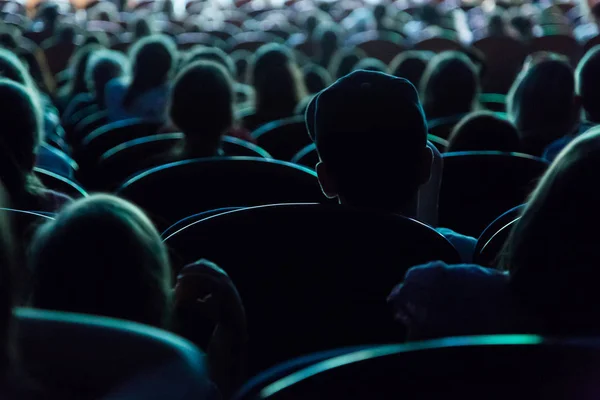 People, children, adults, parents in the theater watching the performance. People in the auditorium looking at the stage. Shooting from the back