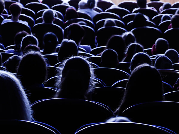 People, children, adults, parents in the theater watching the performance. People in the auditorium looking at the stage. Shooting from the back