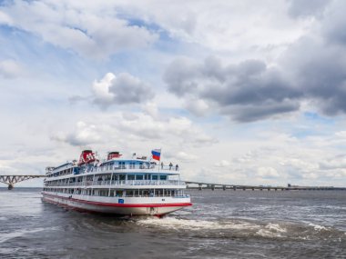 Four-deck passenger ship departs from the pier of the city of Saratov. On the horizon road bridge. The Volga River, Russia. White clouds on a blue sky