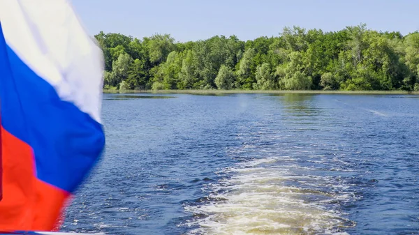 Bandera rusa en la popa del barco. Olas y despertares en el agua. Río o lago en un día de verano. Cielo azul. Vegetación verde en las orillas —  Fotos de Stock