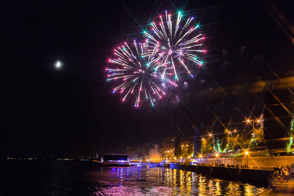 Fireworks over the Volga river in Saratov, Russia. Evening cityscape. People on the street. The moon