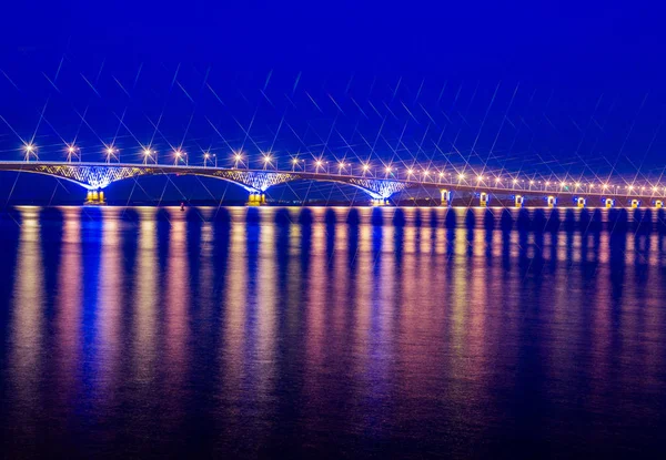 Ponte rodoviária através do rio Volga entre as cidades de Saratov e Engels, Rússia. Paisagem noturna ou noturna. Luzes douradas. O reflexo na água — Fotografia de Stock