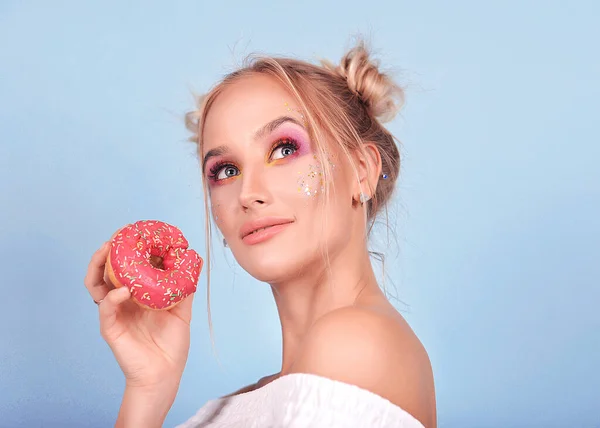 portrait on a blue background a very cute and beautiful girl with an unusual bright makeup loves sweets : sweets, cakes, donuts and macarons . The lady smiles and enjoys life , she is bright and delicious