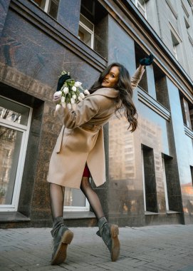 Gülücükler saçan güzel bir kız, gülen çiçeklerle, güneşli havada sokakta portre, lalelerle mutlu kahve içmek, kahkahalar atan bir kitap okumak, bahar mevsiminde makyajlı, ince bir palto giymiş, şık bir mankenle.