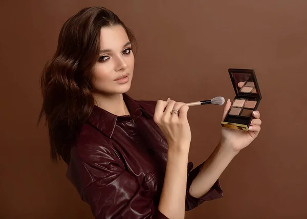 girl makeup artist with brushes, loose powder and eye shadow, on a brown background. The makeup artist is ready to make up. The hair stylist will do the styling and curls, she has combs and Curling iron. Beautiful portrait of a girl smiling sweetly