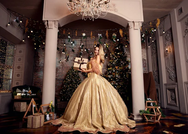 Şatonun lüks şatosu gikarny parlak altın elbisesi içinde korse üzerinde uzun bir tren ve taşlarla süslenmiş güzel prenses kız. Yeni yıl, Noel ağaçları ve hediyeler. Kraliçe çok mutlu ve gülüyor, sana bir hediye veriyor. Karanlık iç mekan, Shine. .