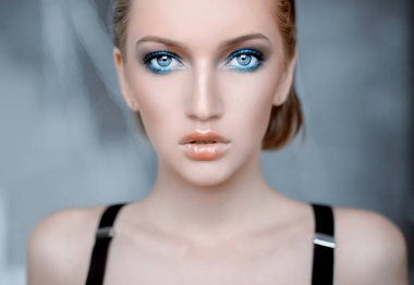 İri mavi gözlü nazik sarışın bir kız. Ev fotoğrafı, portre. Genç ve çekici. Boncuklar, inciler ve tüyler. Sabahlık giymiş bir kız, güneşli bir gün, sıcak ve sıcak bir evde. Alışılmadık parlak makyaj, gelin buketi, çiçekler, bahar