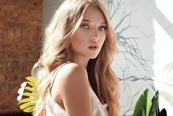 İri mavi gözlü nazik sarışın bir kız. Ev fotoğrafı, portre. Genç ve çekici. Boncuklar, inciler ve tüyler. Sabahlık giymiş bir kız, güneşli bir gün, sıcak ve sıcak bir evde. Alışılmadık parlak makyaj, gelin buketi, çiçekler, bahar