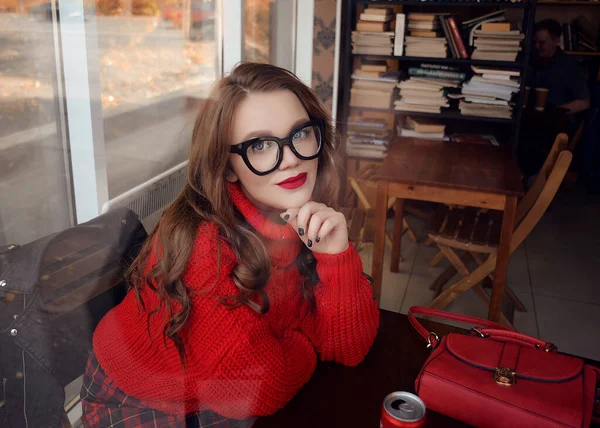 一个穿着红色毛衣和眼镜的漂亮姑娘坐在咖啡店里看书 — 图库照片