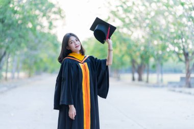 Akademik elbise giyen, mezuniyet şapkası takan genç ve neşeli bir kızın portresi üniversite diplomasını üniversite kampüsünde ilk gününde kutluyor..