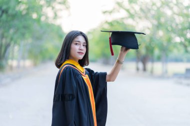 Akademik elbise giyen, mezuniyet şapkası takan genç ve neşeli bir kızın portresi üniversite diplomasını üniversite kampüsünde ilk gününde kutluyor..
