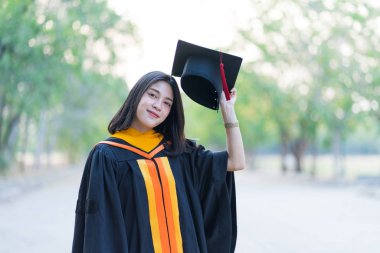 Akademik elbise giyen, mezuniyet şapkası takan genç ve neşeli bir kızın portresi üniversite diplomasını üniversite kampüsünde ilk gününde kutluyor..