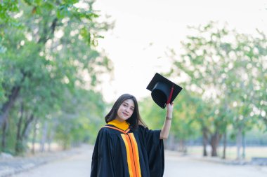Akademik elbise giyen, mezuniyet şapkası takan genç ve neşeli bir kızın portresi üniversite diplomasını üniversite kampüsünde ilk gününde kutluyor..