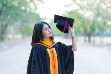 Akademik elbise giyen, mezuniyet şapkası takan genç ve neşeli bir kızın portresi üniversite diplomasını üniversite kampüsünde ilk gününde kutluyor..