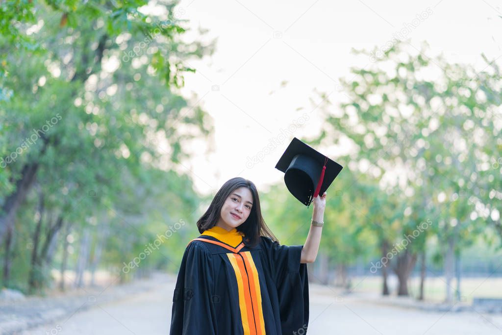 Portrait of a young cheerful female graduate wearing academic gown holding graduate cap celebrates her university degree in commence day in the college campus.