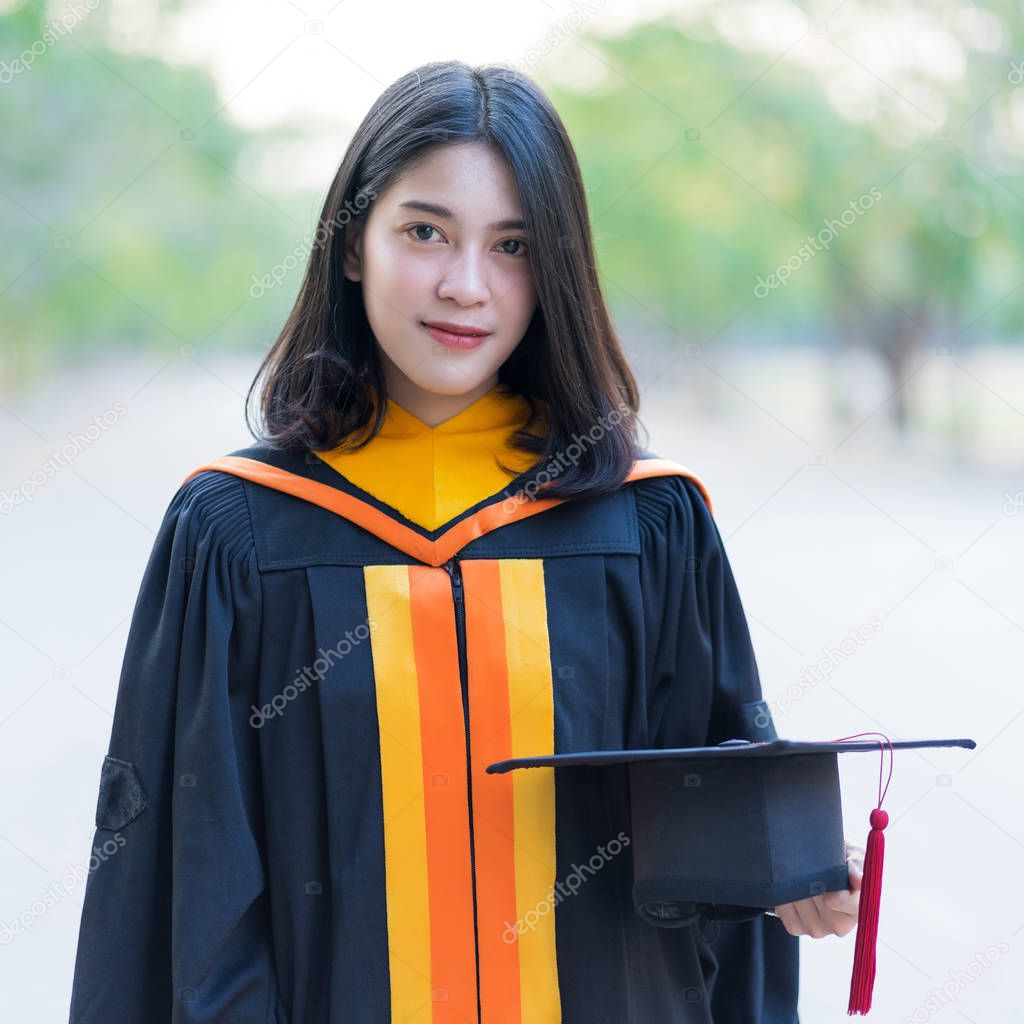 Close up portrait of a young cheerful female graduate wearing academic gown holding graduate cap celebrate her university degree in commence day in the college campus.