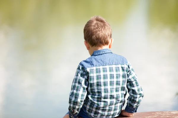 Niño ofendido se volvió se sienta en el banco frente al lago y mira a la distancia —  Fotos de Stock